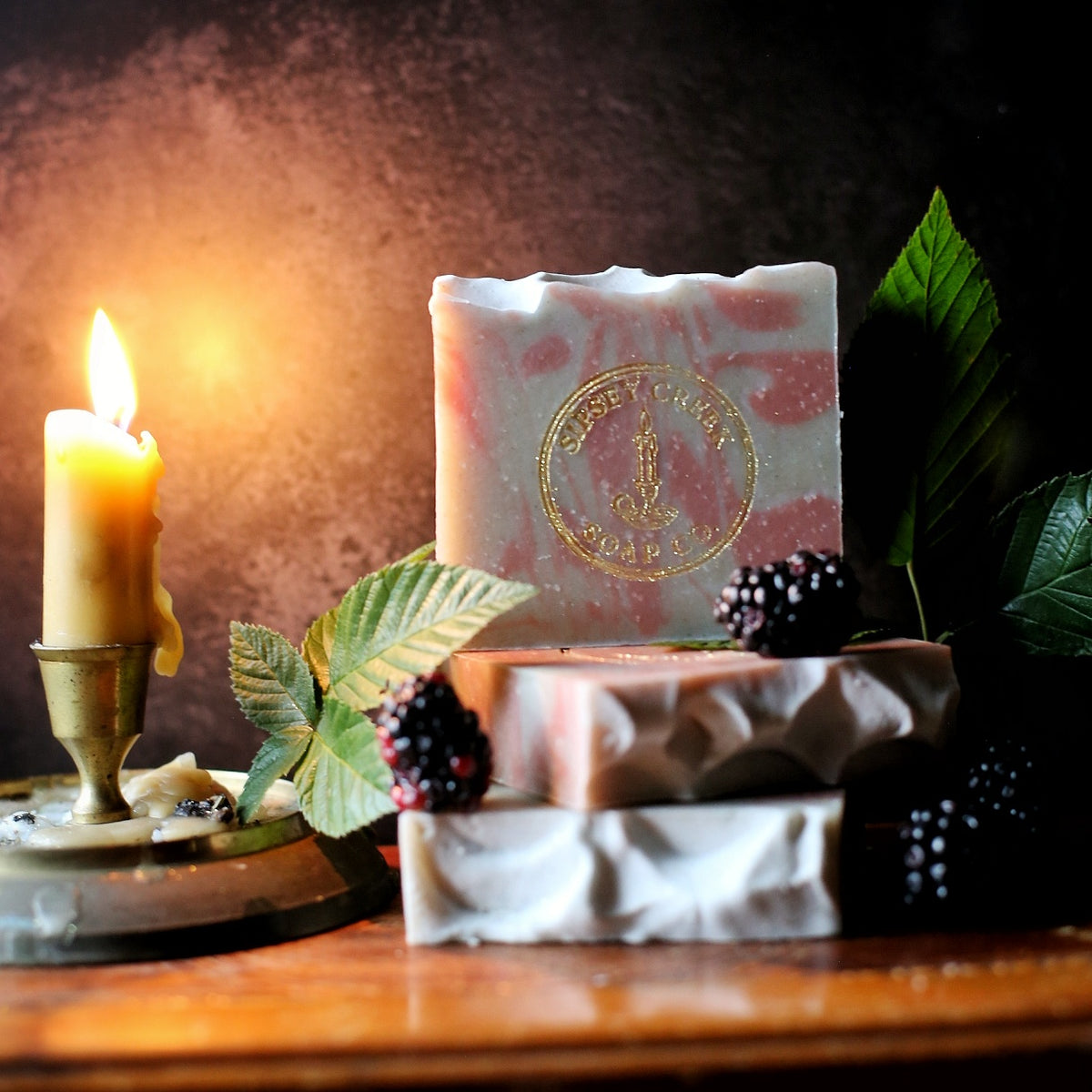 Pink and gray Midnight Bramble soap bars with a gold Sipsey Creek stamp, beside blackberries, leaves, and a lit candle.