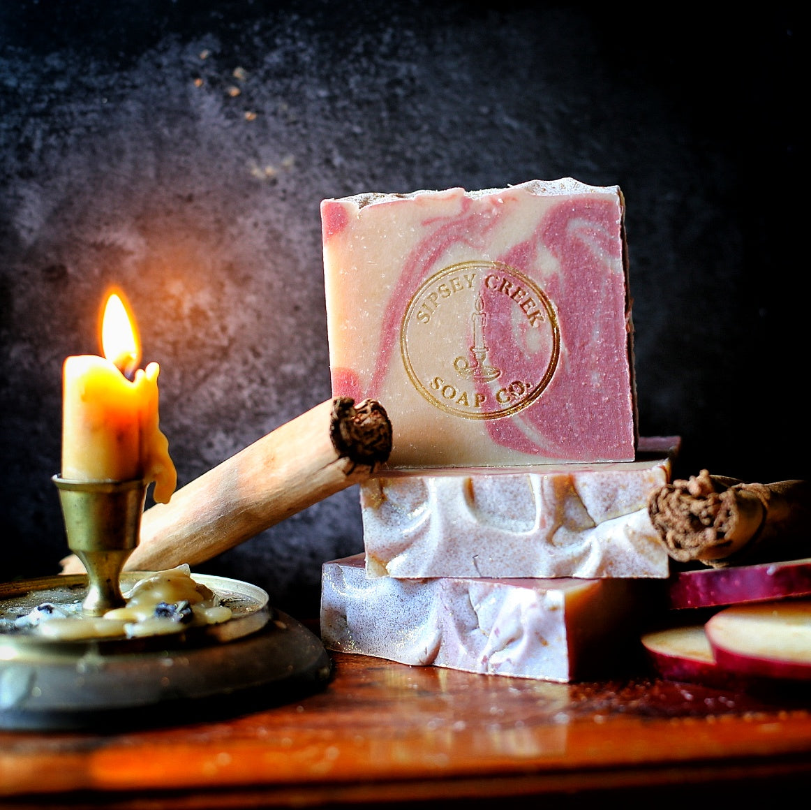 Pink and cream Simmer-Pot soap bars with a gold Sipsey Creek stamp, beside cinnamon sticks, apple slices, and a lit candle.
