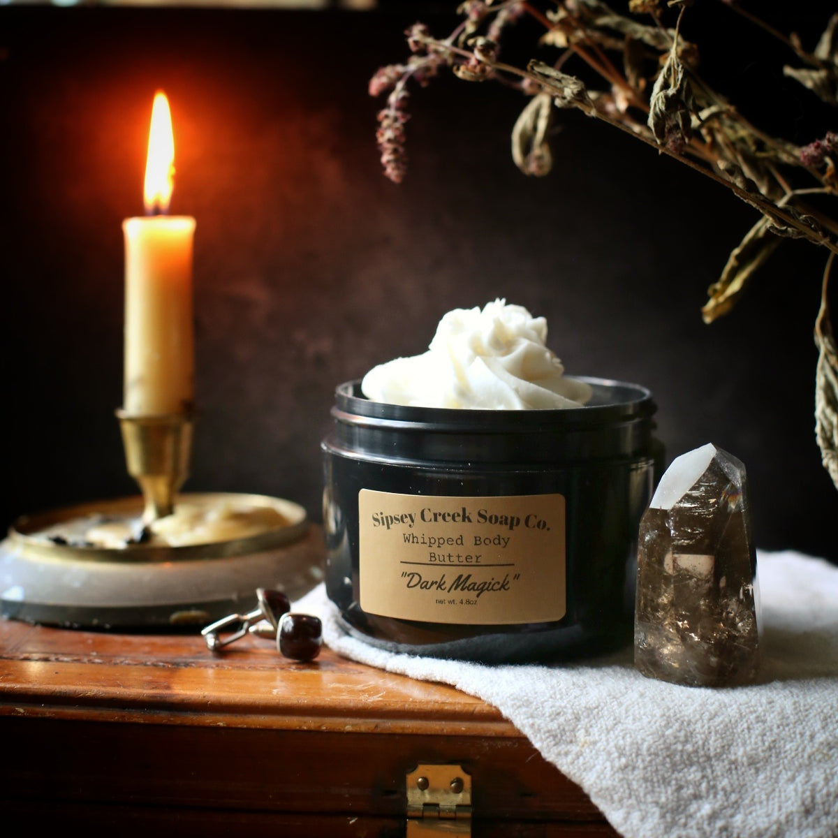 Open black jar of Dark Magick whipped body butter with a gold label, beside a lit candle and smoky crystal on a white towel.