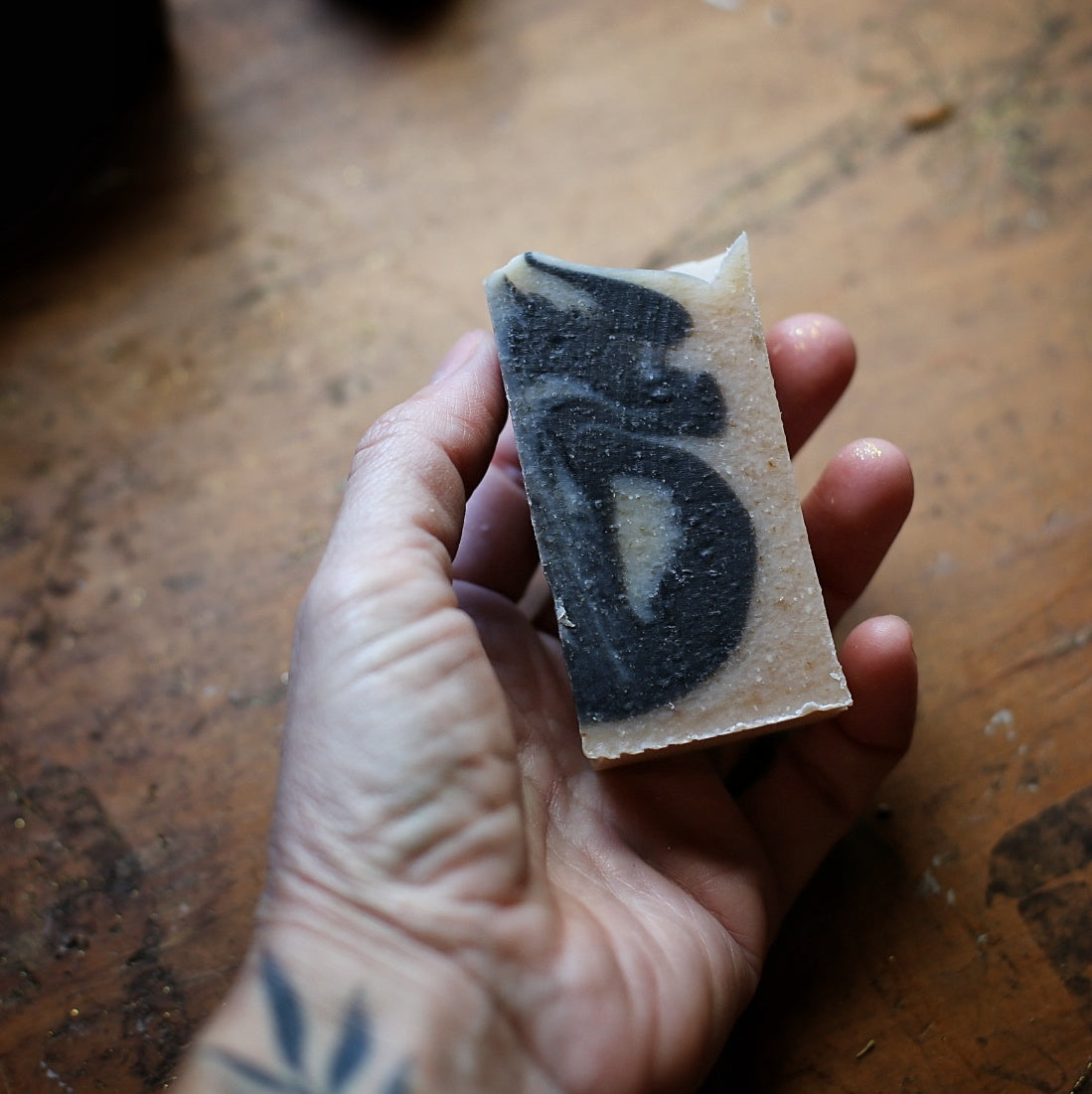 Hand holding a narrow Papa’s Pipe soap bar with dark gray swirls through a cream base, above a wooden tabletop.
