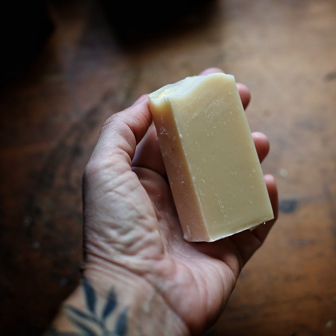 Hand holding a narrow cream-colored Moonlit Laurel soap bar above a wooden tabletop.