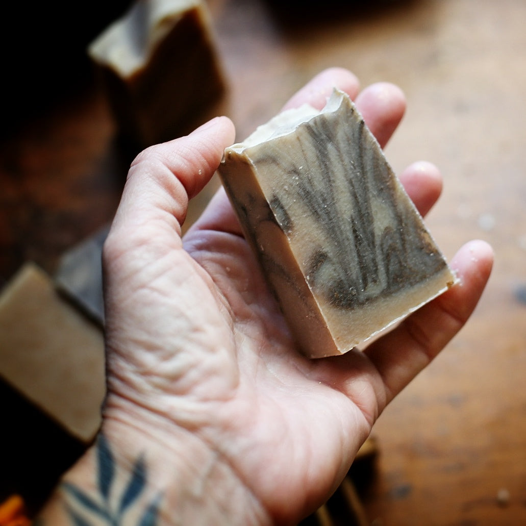 Hand holding a narrow Palo Santo & Vanilla soap bar with gray swirls through a cream base, above a wooden tabletop.