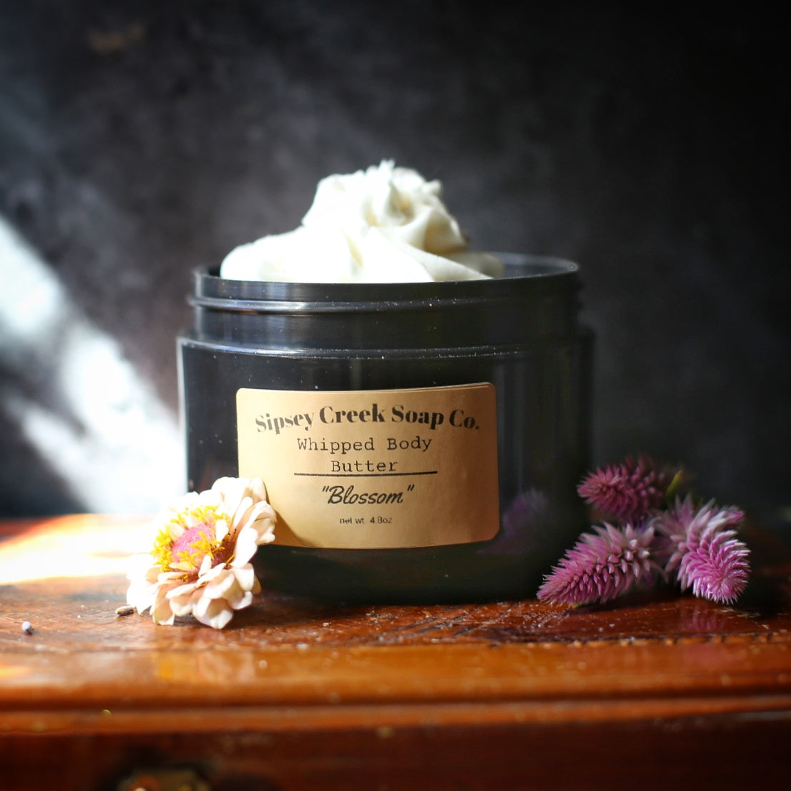 Open black jar of Blossom whipped body butter with a gold label, beside pink and purple flowers on a wooden surface