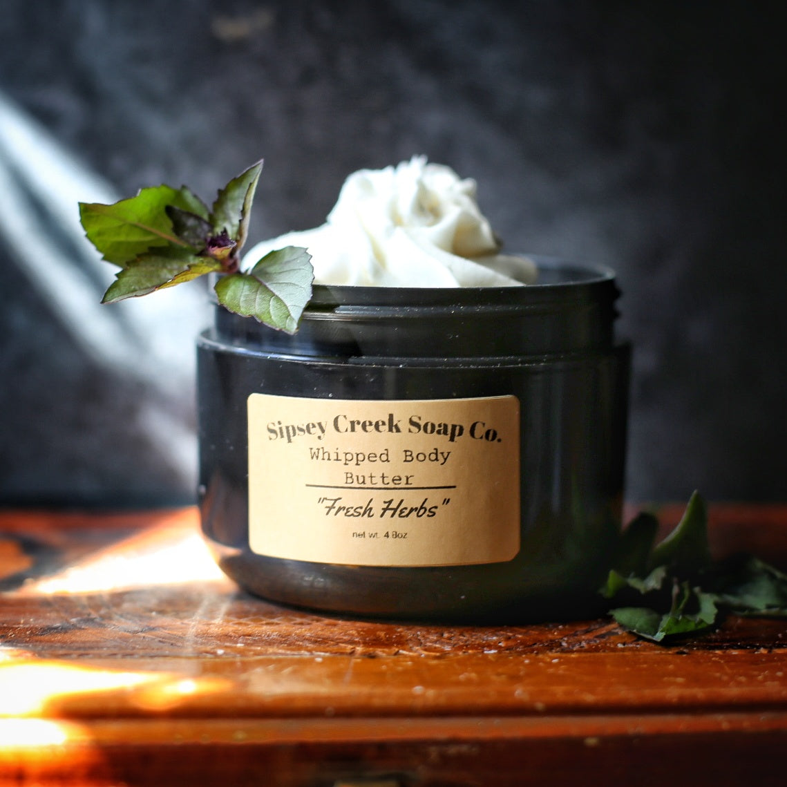 Open black jar of Fresh Herbs whipped body butter with a gold label, styled with leafy herb sprigs on wood