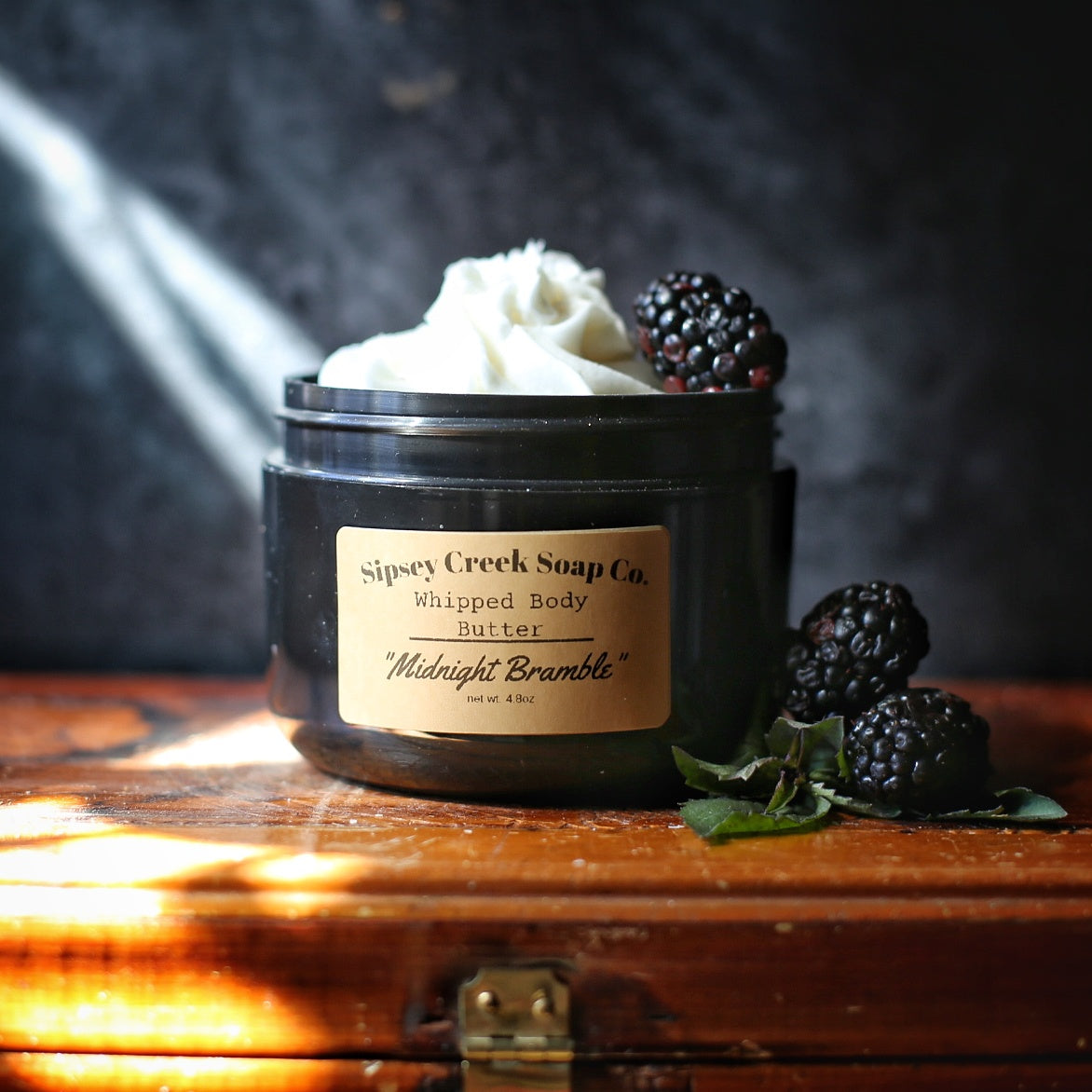 Open black jar of Midnight Bramble whipped body butter with a gold label, styled with blackberries and green leaves.