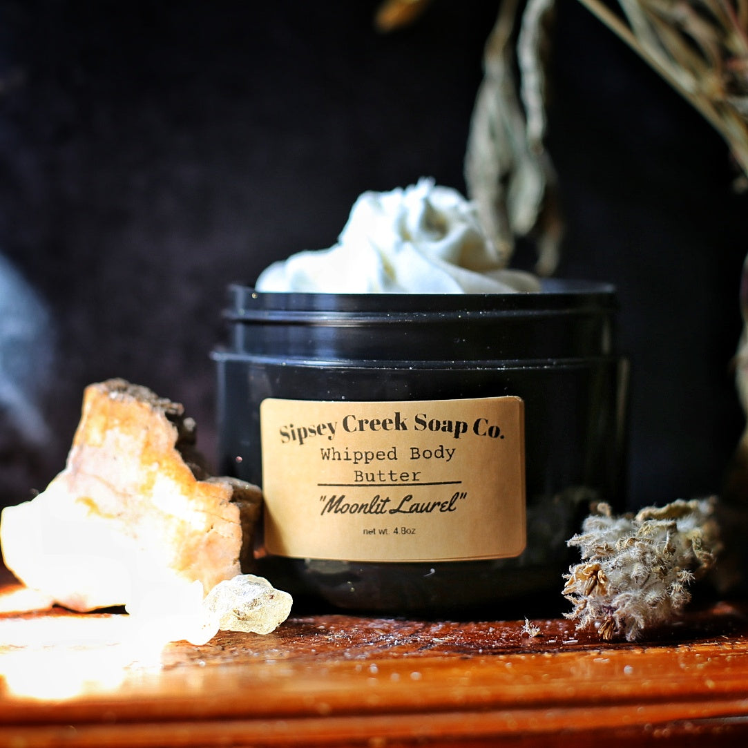 Open black jar of Moonlit Laurel whipped body butter with a gold label, beside pale crystals and dried flowers on wood
