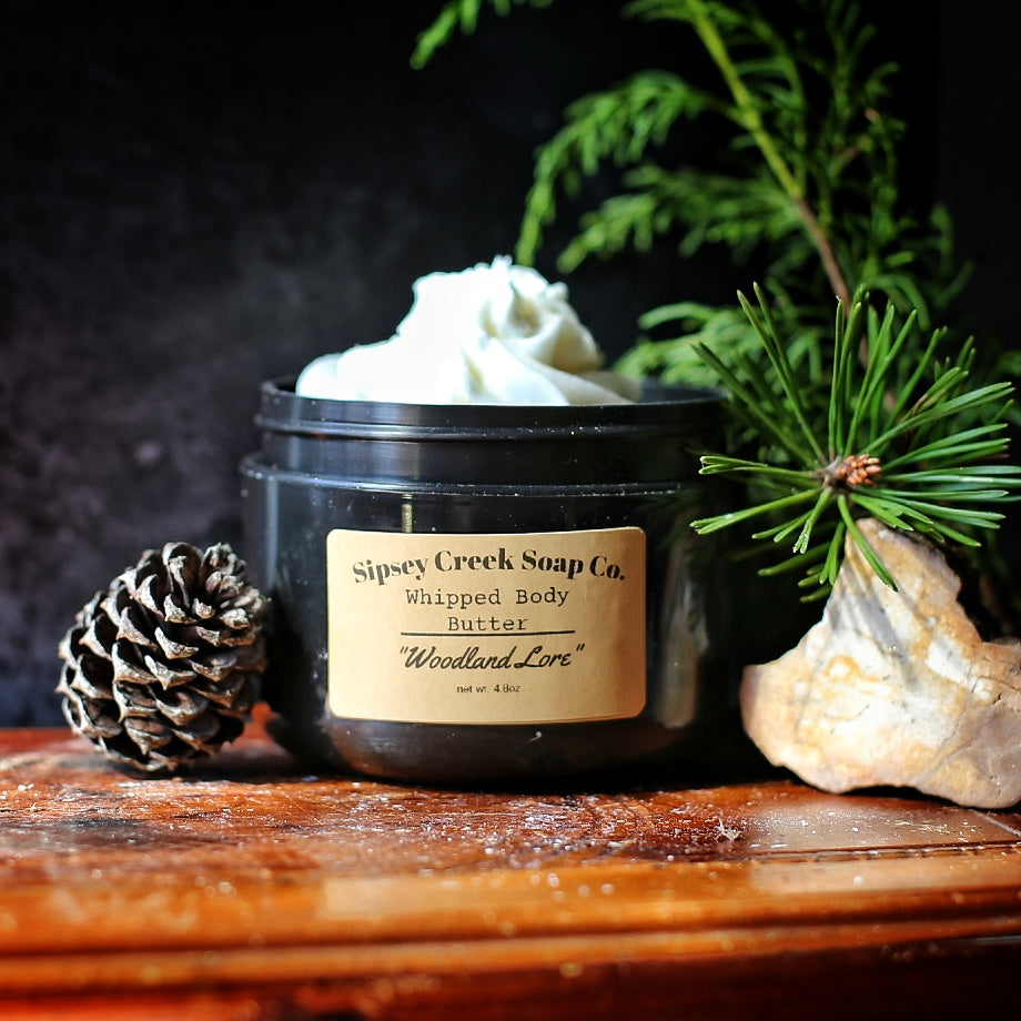 Open black jar of Woodland Lore whipped body butter with a gold label, beside a pine cone, evergreen sprigs, and a pale rock