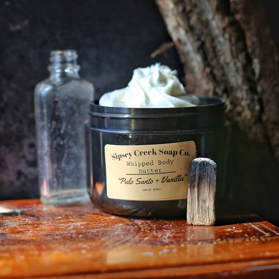 Open black jar of Palo Santo & Vanilla whipped body butter beside a charred wood stick and clear glass bottle.
