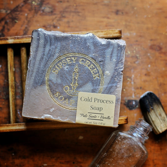 Wrapped Palo Santo & Vanilla soap with its front label, on a wooden soap dish beside a charred wood stick and glass bottle.