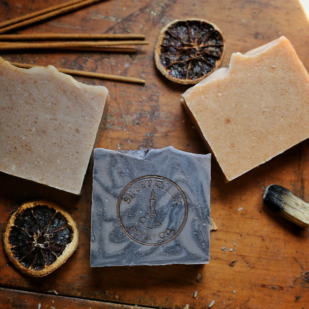 Three Apothecary Collection soap bars in orange, peach, and taupe with textured tops, arranged on draped cream fabric.