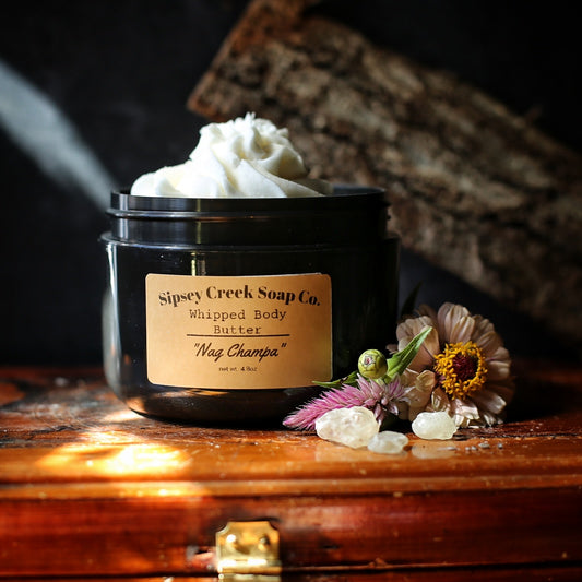 Open black jar of Nag Champa whipped body butter with a gold label, beside pink flowers and pale crystals on wood