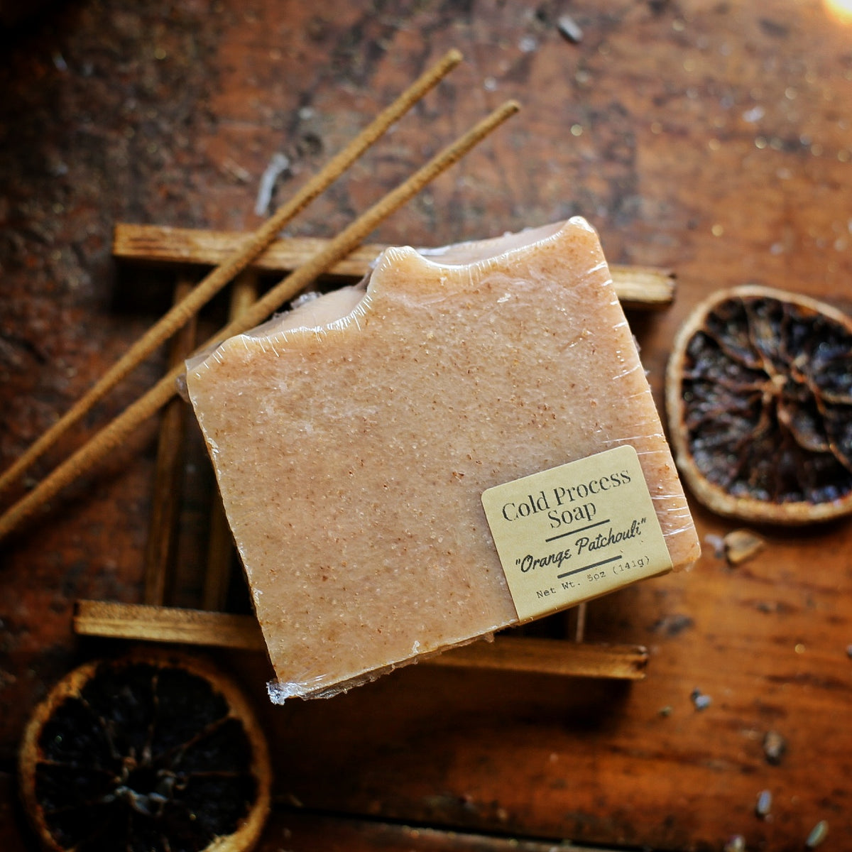 Wrapped tan Orange Patchouli soap with its front label, on a wooden soap dish beside dried orange slices and incense sticks.