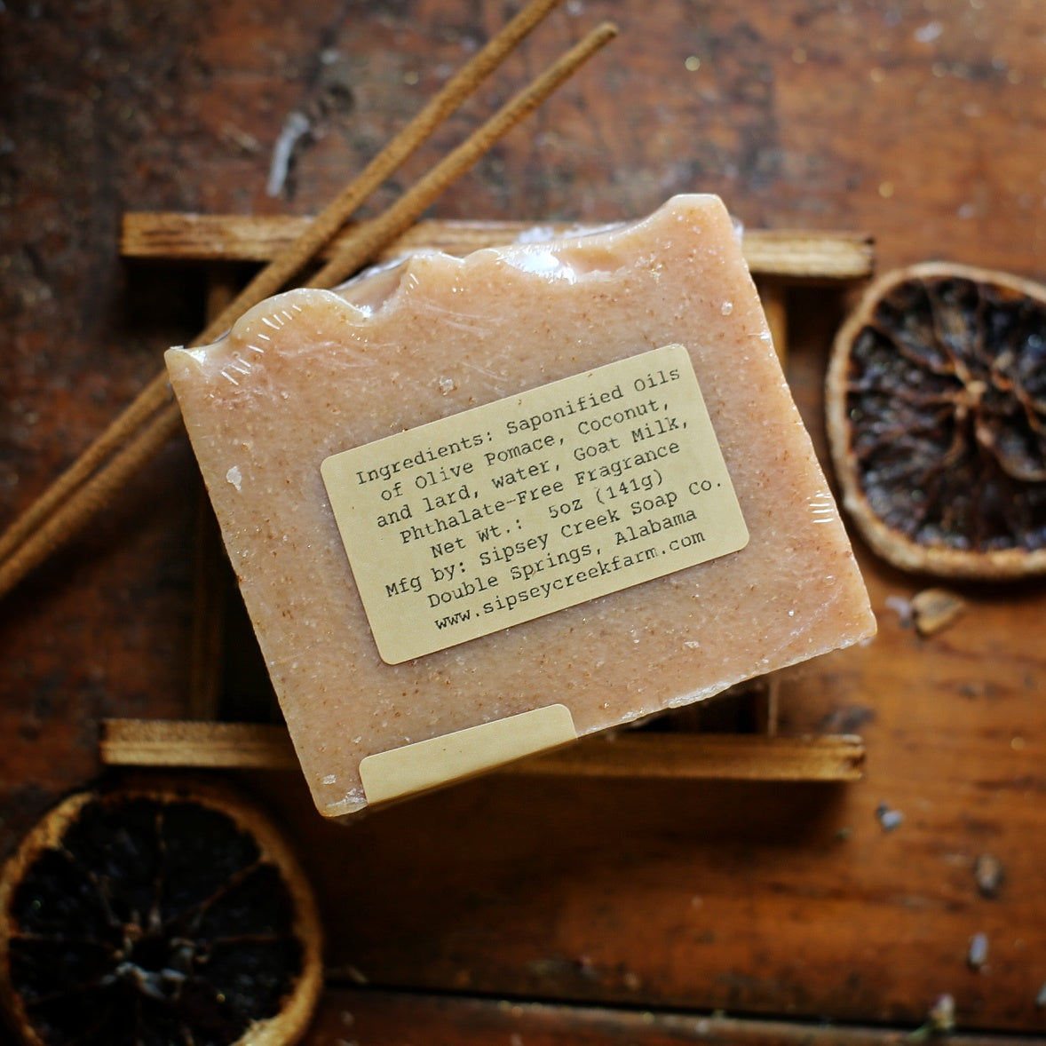 Back of wrapped Orange Patchouli soap showing its ingredient label, on a wooden dish beside dried orange slices.