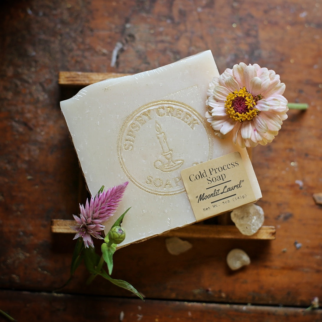 Wrapped cream Moonlit Laurel soap with a gold Sipsey Creek logo and front label, on a wooden dish beside flowers.