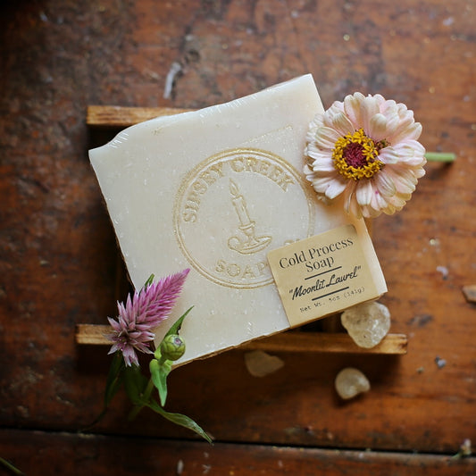 Wrapped cream Moonlit Laurel soap with a gold Sipsey Creek logo and front label, on a wooden dish beside flowers.