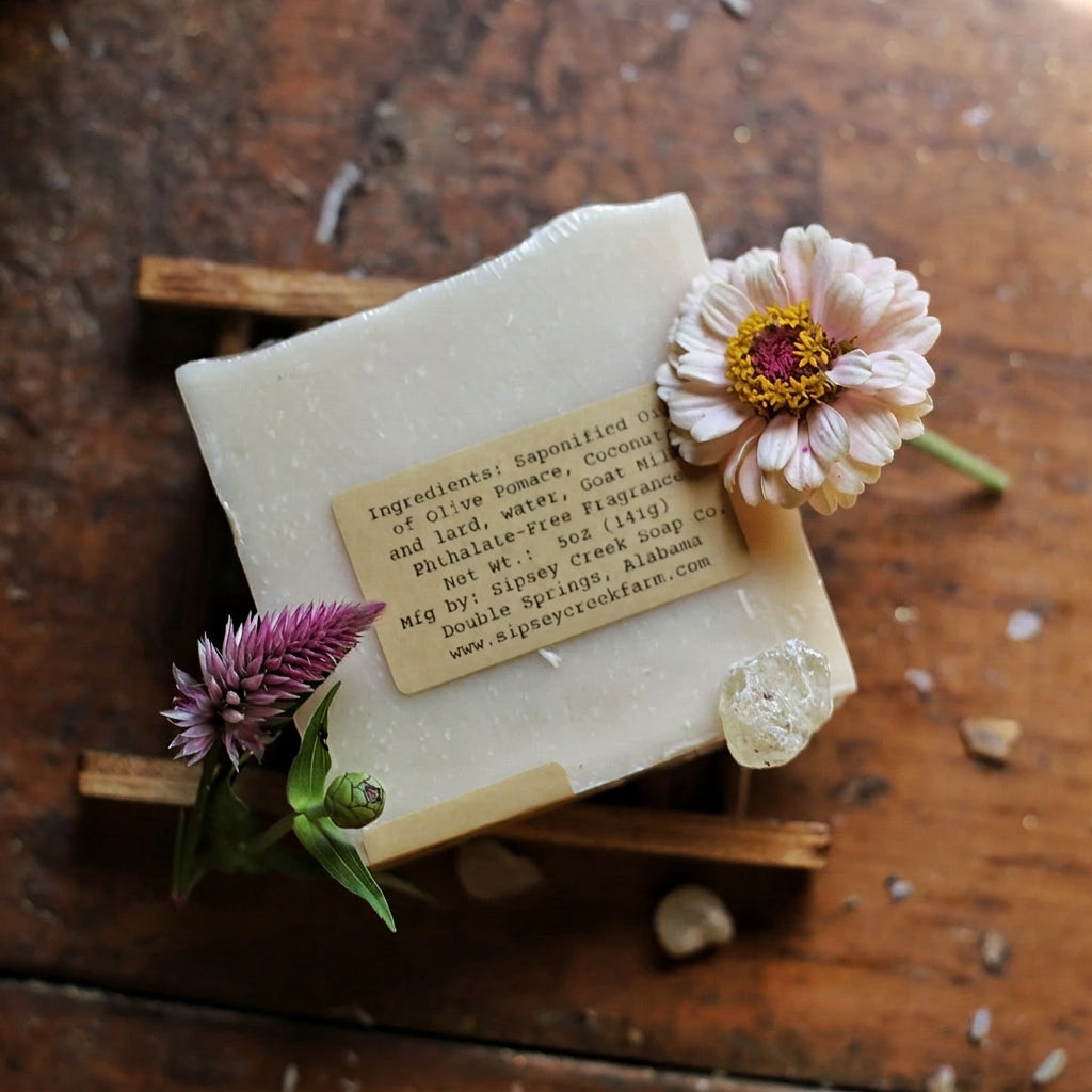 Back of wrapped Moonlit Laurel soap showing its ingredient label, on a wooden dish beside pink and purple flowers