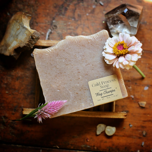 Wrapped tan Nag Champa soap with its front label, resting on a wooden soap dish beside pink flowers and crystals.