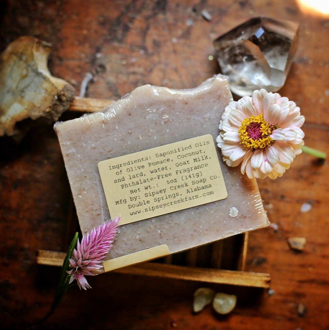Back of wrapped Nag Champa soap showing its ingredient and 5-ounce weight label, on a wooden dish beside pink flowers.