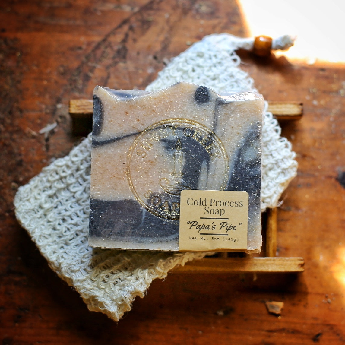 Wrapped Papa’s Pipe soap with cream and dark gray swirls and a front label, on a knitted cloth over a wooden soap dish.