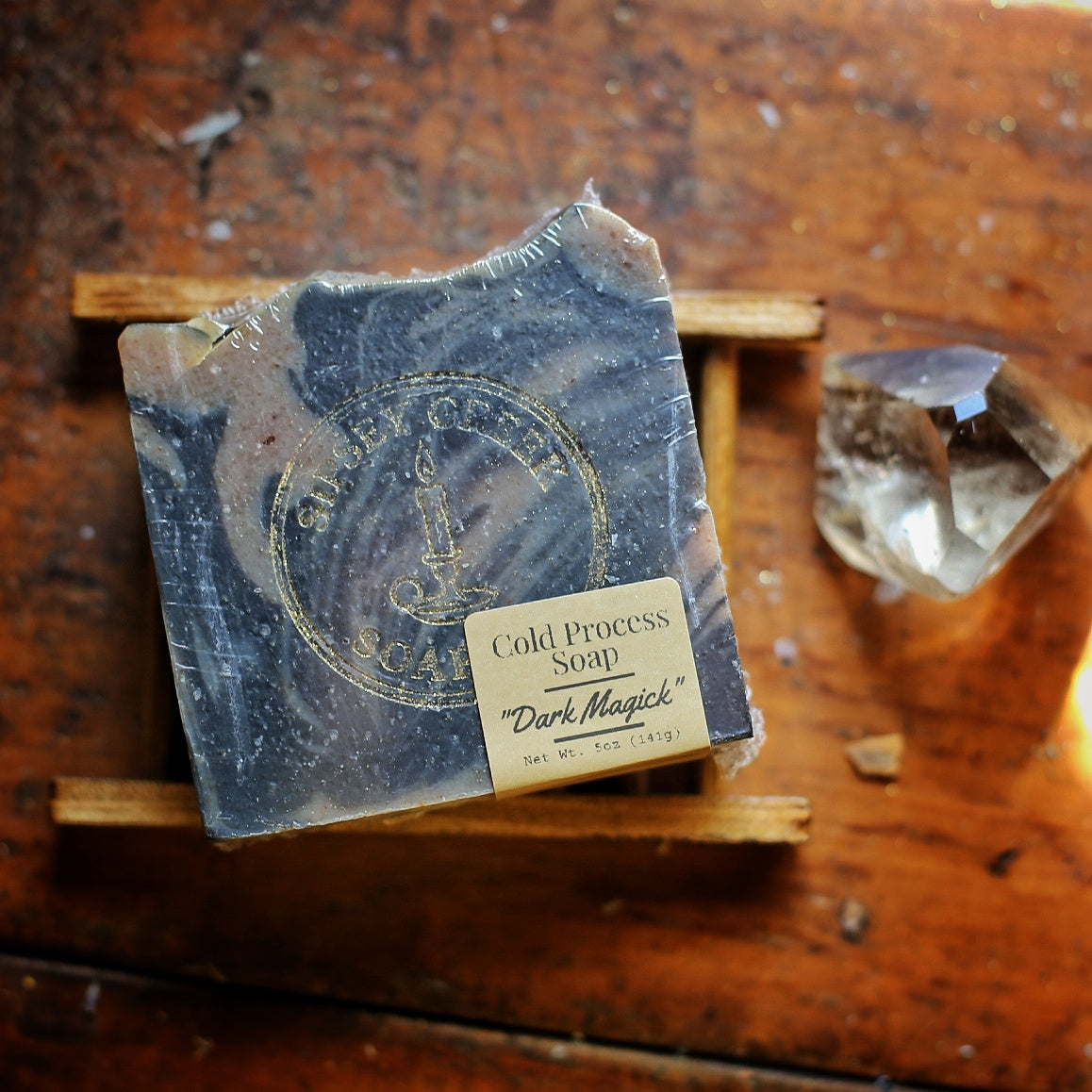 Wrapped Dark Magick soap with dark gray and cream swirls, a gold Sipsey Creek logo, and front label, on a wooden soap dish nest to a clear crystal.