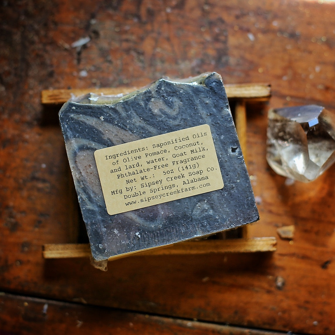 Back of wrapped Dark Magick soap showing its ingredient and 5-ounce weight label, on a wooden dish beside a clear crystal