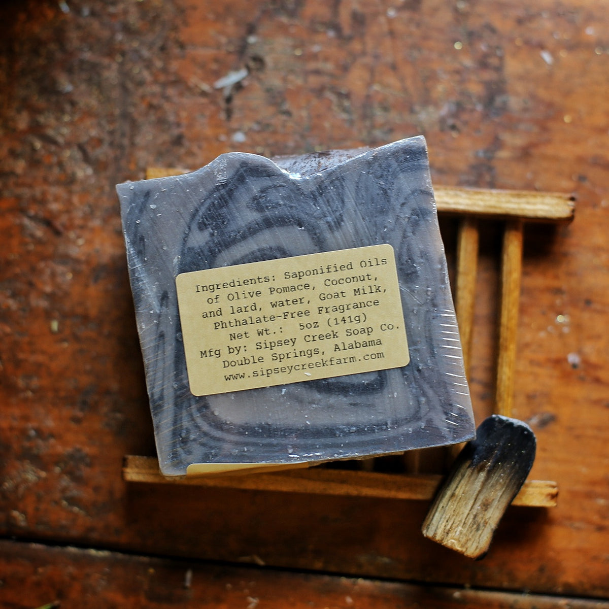 Back of wrapped Palo Santo soap showing its ingredient and 5-ounce weight label, on a wooden dish beside a charred wood stick.
