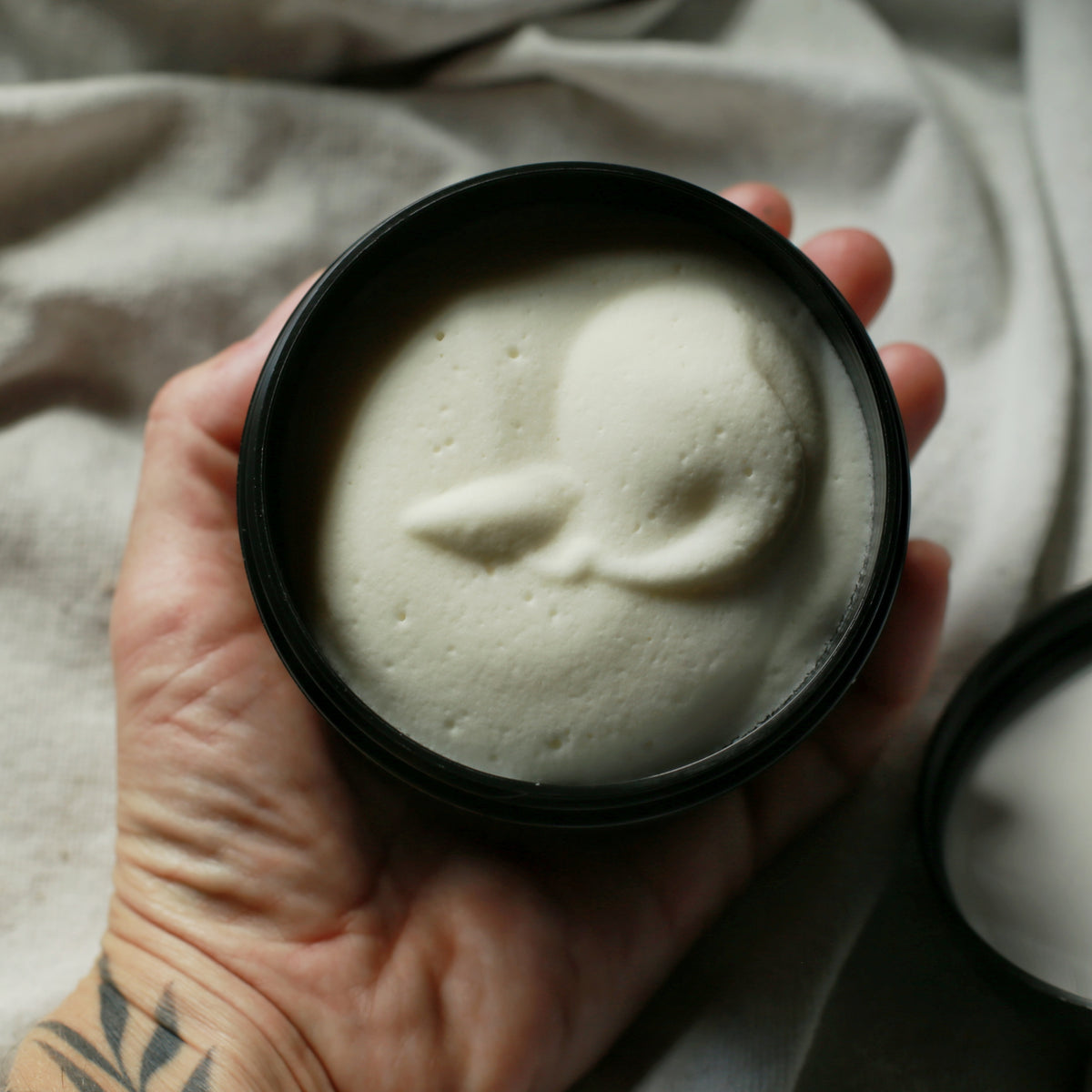 A hand holds a round black tin filled with a thick, off-white cream or balm, with the lid resting beside it on draped fabric.