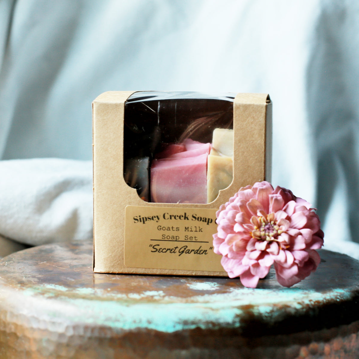 A boxed soap set from Sipsey Creek Soap on a rustic wooden surface, with a pink flower prop in front. The kraft paper box has a clear window showing pink and cream bars inside.