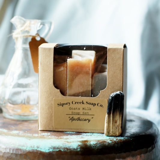 A boxed soap set from Sipsey Creek Soap Co. labeled 'Apothecary' with 'Goats Milk Soap Set' on the front, shown on a rustic surface with a glass bottle in the background.