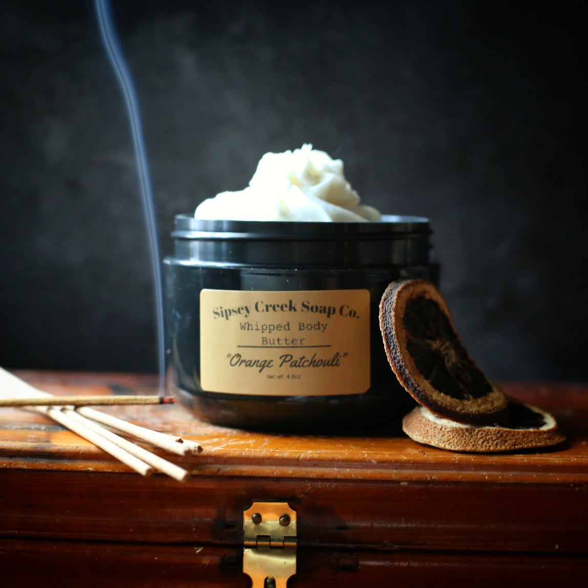 A black jar of Sipsey Creek Soap Co. Whipped Body Butter in the Orange Patchouli scent, filled with white whipped cream and topped with a small sprig of fresh rosemary.