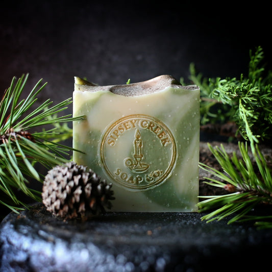 Green and cream Woodland Lore goat’s milk soap with an embossed Sipsey Creek logo, on a cast iron plate, surrounded by pine sprigs and a pine cone.