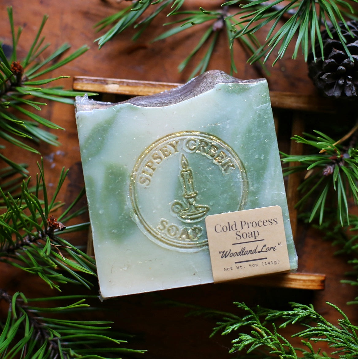 Shrink Wrapped Woodland Lore goat’s milk soap with its front label, resting on a wooden soap dish among evergreen sprigs.