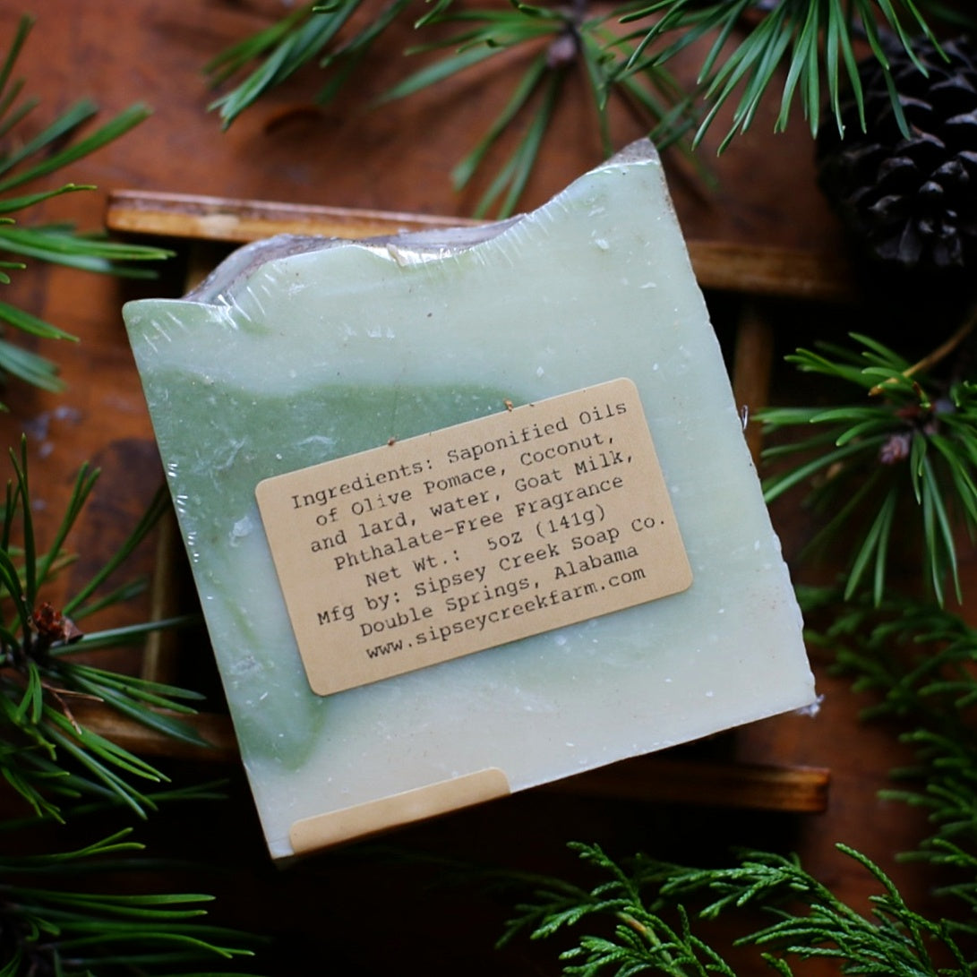 Back of wrapped Woodland Lore goat’s milk soap showing the ingredient and 5-ounce weight label among evergreen sprigs.