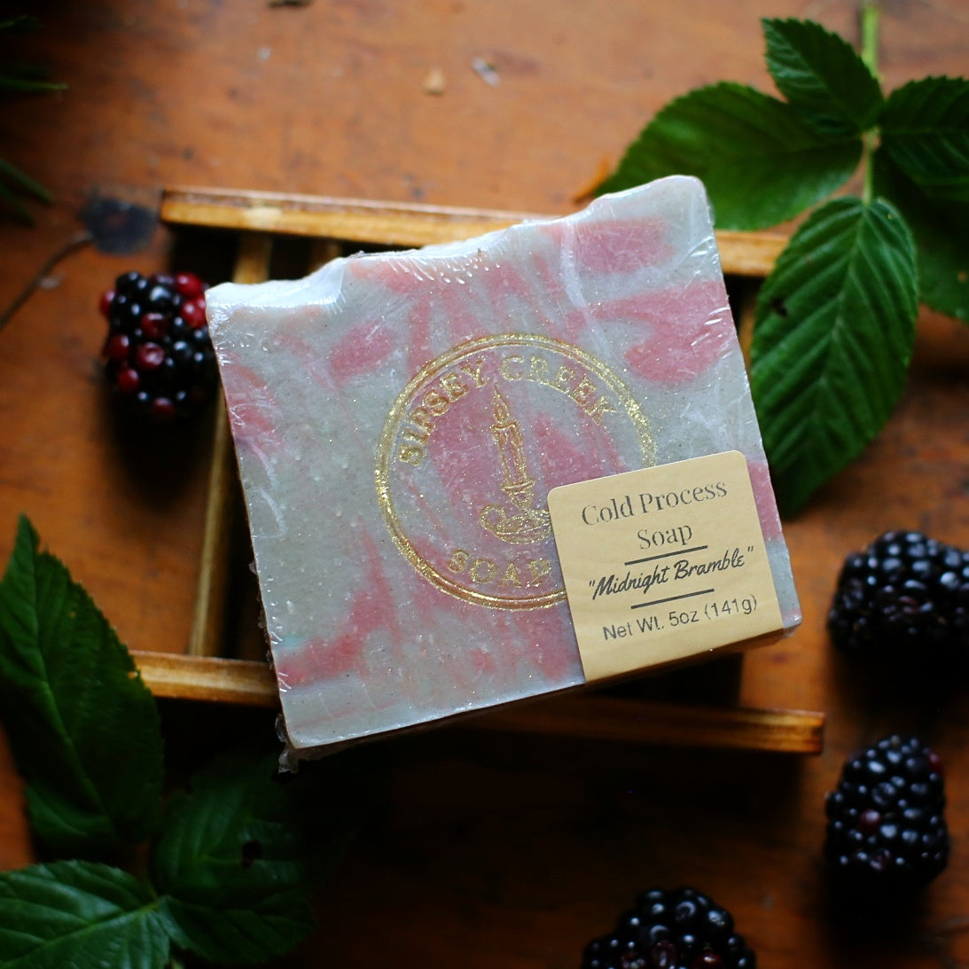 Wrapped Midnight Bramble soap with pink and gray swirls and a 5-ounce front label, on a wooden dish beside blackberries.