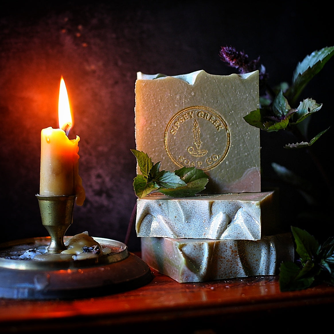 Stacked green and cream Fresh Herbs soap bars with a gold Sipsey Creek stamp, beside leafy herbs and a lit candle.