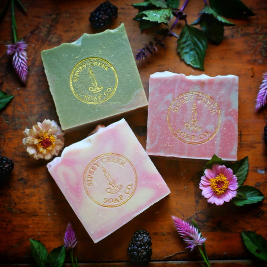 Three Secret Garden soap bars in green, pink, and cream with gold Sipsey Creek stamps, among flowers, herbs, and blackberries.
