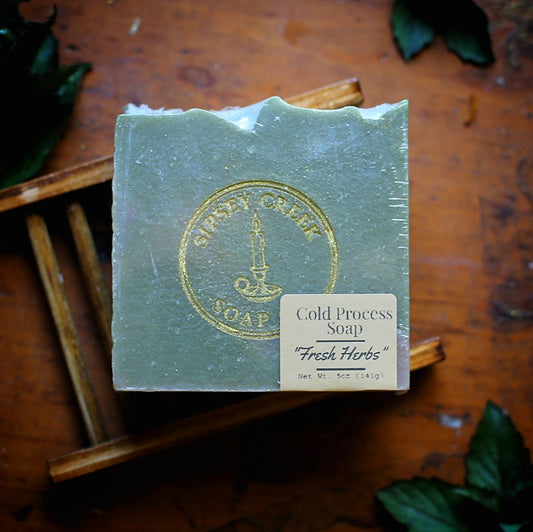 Wrapped green Fresh Herbs soap with a gold Sipsey Creek stamp and 5-ounce front label, resting on a wooden soap dish.