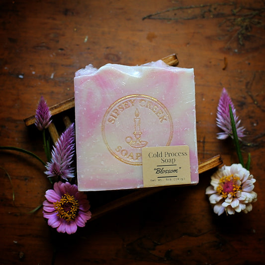Wrapped pink and cream Blossom soap with its front label, resting on a wooden soap dish surrounded by purple and white flowers.