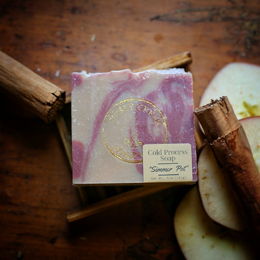 Wrapped pink and cream Simmer-Pot soap with its front label, on a wooden soap dish beside cinnamon sticks and apple slices.
