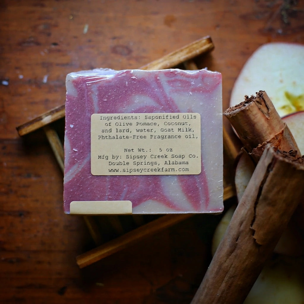 Back of wrapped Simmer-Pot soap showing its ingredient label, on a wooden dish beside cinnamon sticks and apple slices.