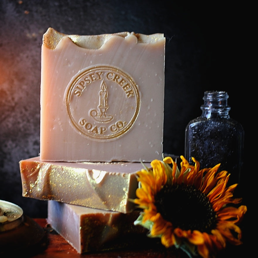 Stacked Sipsey Gold soap bars with gold accents and an embossed Sipsey Creek logo, beside a sunflower and glass bottle.