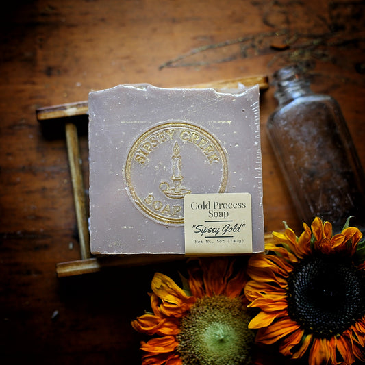 Wrapped Sipsey Gold soap with its 5-ounce front label and gold logo, on a wooden soap dish beside sunflowers.