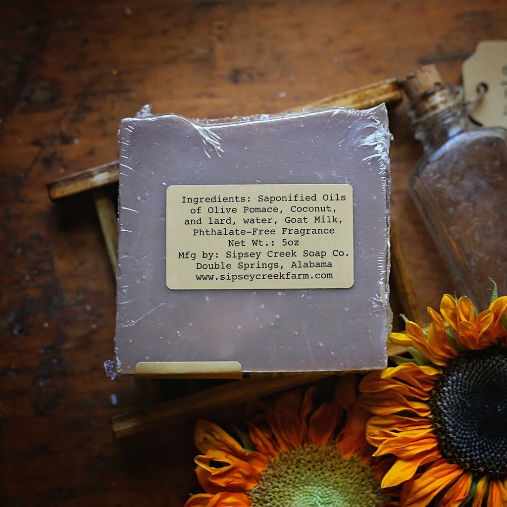 Back of wrapped Sipsey Gold soap showing its ingredient label, on a wooden soap dish beside sunflowers and a glass bottle.